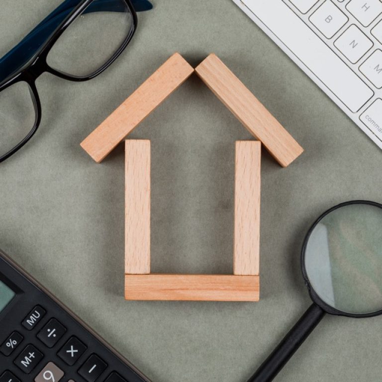 Real estate concept with house made of wooden blocks, glasses, magnifying glass, keyboards on grey background close-up.